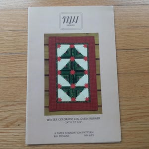 May include: A paper foundation pattern for a Winter Colorado Log Cabin Runner, featuring a rectangular design with green, red, and white geometric shapes. The pattern measures 14" x 22 1/4" and is from MH Designs.