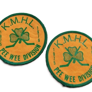 May include: Two circular patches with a gold background and green edging. The patches display the text "K.M.H.L. 1975-1976 PEE WEE DIVISION" around a green shamrock and hockey sticks.