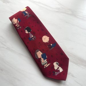 Peut inclure: Une cravate bordeaux ornée d'un motif répétitif représentant les personnages des Peanuts, dont Charlie Brown, Lucy et Snoopy, entremêlés de lettres noires dispersées. Les personnages sont représentés dans leur style illustré classique.