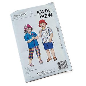 May include: A Kwik Sew pattern envelope 3079 for children's clothing. The illustration shows two figures wearing bucket hats, one in a blue shirt with plaid trousers and the other in a white shirt with a sailboat print and blue shorts. Includes text.