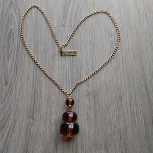 May include: A gold-toned chain necklace with a pendant featuring three stacked, dark amber-colored beads. The chain is arranged in a heart shape. A small rectangular tag is attached to the chain.