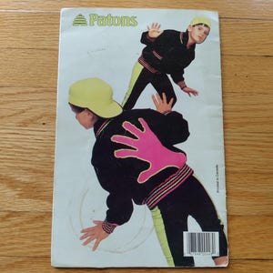 May include: A rectangular Patons knitting pattern booklet featuring a graphic of two figures wearing black jackets with neon pink hand prints on the back, neon yellow caps, and black pants with neon yellow side stripes. The back cover includes a barcode.