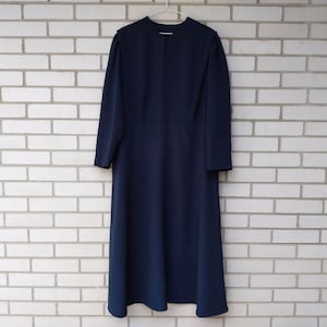 May include: A navy blue, long-sleeved dress hangs against a white brick wall. The dress has a simple design with a round neckline and a flowing skirt. The dress is made of a smooth, solid fabric. The dress is a classic style.