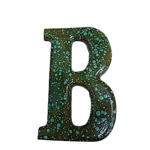 May include: A decorative letter "B" in a rustic style. The letter is dark green with a speckled turquoise pattern. The design has a distressed, aged appearance, suitable for home decor.