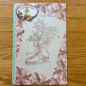 May include: A rectangular craft pattern booklet titled Vintage Ice Skate by Crab-Apple Hill. The cover features a central illustration of an ice skate adorned with a pink bow, candy canes, and holly, set against a pink and white floral quilt pattern.