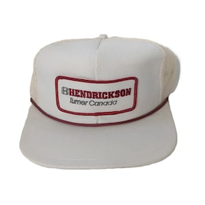 May include: White trucker hat with a mesh back and a curved brim. The front features a rectangular patch with the text "HENDRICKSON Turner Canada" in red and white. A red rope detail accents the brim.