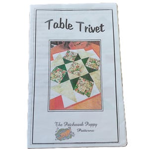 May include: A white paper pattern booklet titled Table Trivet with a central color photograph showing a square quilted trivet featuring green, white, and floral fabric patches. The bottom includes the text The Patchwork Puppy Patterns.