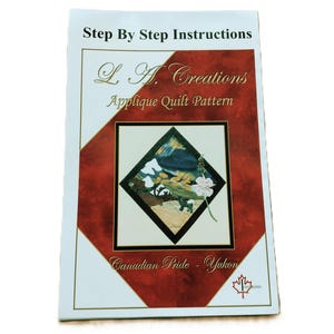 May include: A printed instructional booklet titled L.A. Creations Applique Quilt Pattern, Canadian Pride - Yukon. The cover features a diamond-shaped illustration of a landscape with a white flower and a small animal, set against a deep red background.