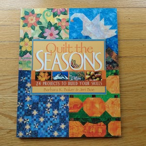 May include: A softcover book titled Quilt the Seasons: 24 Projects to Build Your Skills by Barbara K. Baker and Jeri Boe. The cover features a colorful collage of quilt patterns including floral motifs, a white bird, stars, and orange pumpkins.