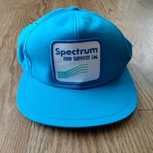Peut inclure: Casquette de baseball bleu vif avec une visière incurvée. Elle présente un écusson carré blanc avec le texte "Spectrum FEED SERVICES Ltd." en bleu et un motif de vagues vertes. La casquette est faite d'un tissu résistant.