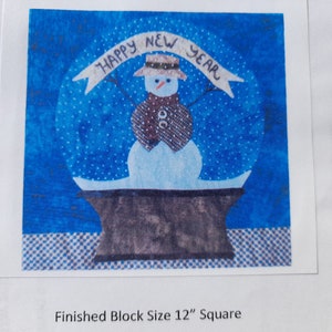 May include: A square fabric quilt block featuring a snowman in a blue snow globe with the text HAPPY NEW YEAR on a banner. The snowman wears a brown hat and scarf. The block measures 12 inches square as indicated by the printed text.