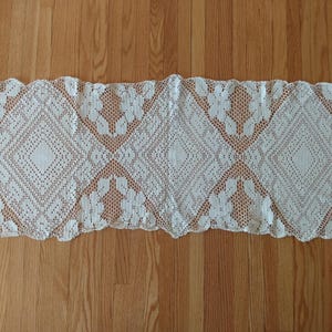 May include: A rectangular off-white crochet table runner featuring an intricate openwork pattern. The design consists of repeating diamond-shaped motifs interspersed with floral elements, finished with a scalloped edge along the entire perimeter.