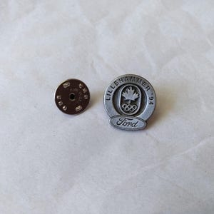 May include: A circular silver-toned metal lapel pin featuring the text LILLEHAMMER '94, a maple leaf, and the Olympic rings, with the Ford logo at the bottom. The pin includes a separate circular metal clutch backing with a textured edge.
