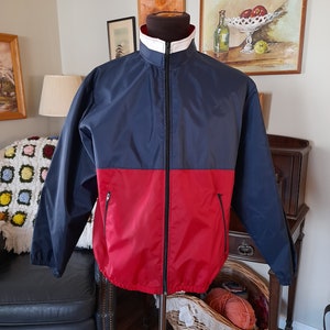 May include: A two-tone jacket in navy blue and red, displayed on a mannequin. The jacket has a white collar and a full-length zipper. Two pockets are visible on the front. The jacket is made of a lightweight material.