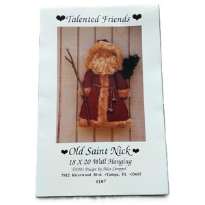 May include: A craft pattern sheet titled Talented Friends and Old Saint Nick 18 X 20 Wall Hanging. The image shows a Santa figure in a deep red coat with tan fur trim, holding a brown stick and a small green tree against a light wood background.