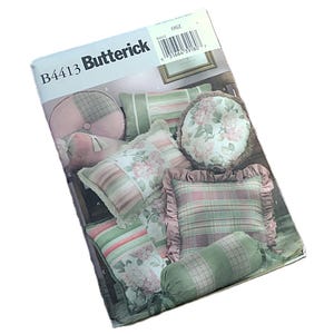 May include: A Butterick sewing pattern, B4413, featuring various pillow designs. The pillows showcase floral and plaid patterns in shades of pink, green, and white. The pattern includes round, square, and bolster-style pillows.