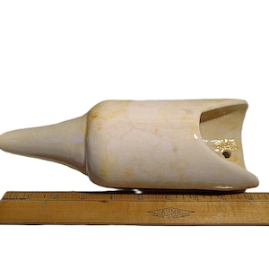 May include: Cream-coloured ceramic object with a spout and a curved opening. The object is approximately 20 cm long and is placed next to a wooden ruler.