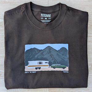 May include: Dark brown t-shirt with a graphic of a camper, car, and mountains. The graphic has the text "SHE'LL BE RIGHT" and "TEEDOG". The shirt is folded on a light brown wooden surface.