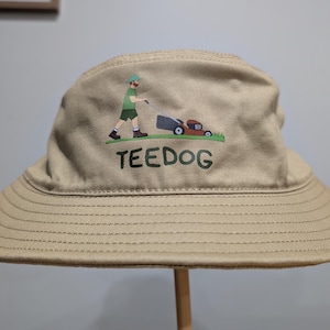 Peut inclure: Chapeau de pêcheur beige avec un bord large, orné d'une illustration d'une personne tondant la pelouse, avec le mot "TEEDOG" en vert. Le chapeau est en tissu résistant, idéal pour les activités en plein air.