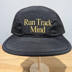 Puede incluir: Gorra negra de cinco paneles con visera plana. La gorra presenta las palabras "Run Track Mind" en letras doradas en la parte delantera. El sombrero se exhibe sobre un soporte de madera sobre un fondo neutro.