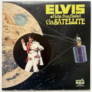 May include: Album cover for "Elvis: Aloha from Hawaii via Satellite." The cover features a circular image of Elvis Presley in a white jumpsuit with red accents, set against a backdrop of Earth and space. The title is in yellow and white.