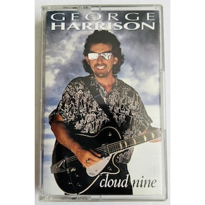 May include: A cassette tape of George Harrison's album "Cloud Nine." The cover features a smiling man wearing sunglasses and holding a black guitar. The man is wearing a black and white patterned shirt. The album title is in white script.