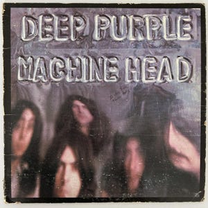 May include: Vintage album cover for Deep Purple's "Machine Head." The album title and band name are embossed in silver lettering. The background features a blurred image of the band members. The cover has a black border and some wear.