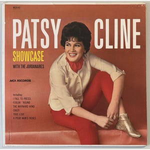 May include: Vintage vinyl record album cover for Patsy Cline's "Showcase" album. The cover features a photo of Patsy Cline, with the album title in large white letters. The album includes songs like "Crazy" and "True Love".