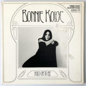 May include: A vintage album cover featuring the artist Bonnie Koloc. The black and white image shows Koloc with her arms crossed. The album title "Hold On To Me" is at the bottom. The cover has decorative framing and text.