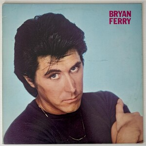 May include: Album cover featuring Bryan Ferry with a serious expression. He wears a black shirt and a silver necklace. The background is a light blue. The text "BRYAN FERRY" is in the upper right corner in pink.