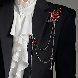 May include: A black blazer with a white ruffled shirt and a red floral brooch with dangling silver chains. The brooch features red flower accents and is attached to the blazer's lapel, adding a touch of elegance.