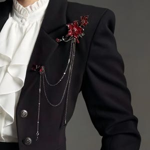 May include: A black blazer adorned with two red floral brooches. The brooches are connected by delicate silver chains. The blazer is worn over a white ruffled shirt.