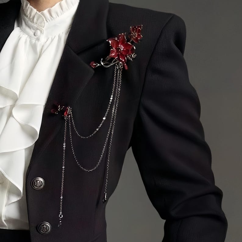 May include: A black blazer adorned with two red floral brooches. The brooches are connected by delicate silver chains. The blazer is worn over a white ruffled shirt.