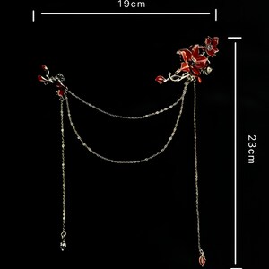 May include: A decorative hair accessory featuring red floral accents and dangling chains. The piece is 19 cm wide and 23 cm long. The design includes silver-toned chains and red flower details, set against a black background.