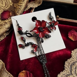 May include: A black metal floral brooch with red enamel flowers and dangling chains. The brooch features a stylized flower design with multiple petals and a dark, gothic aesthetic. The brooch is displayed on a white card against a red velvet background.
