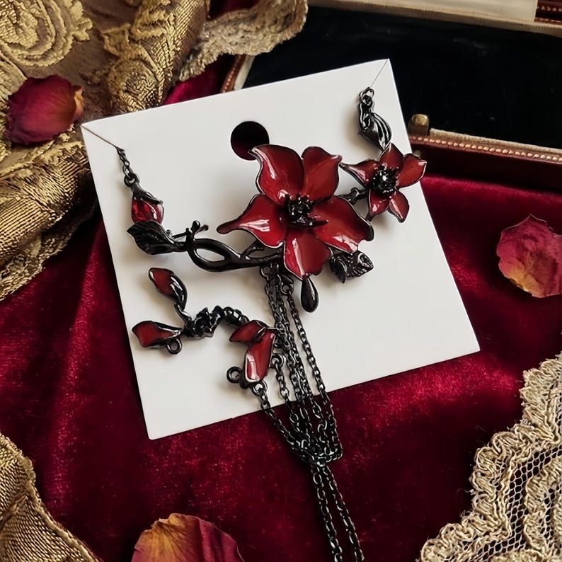 May include: A black metal floral brooch with red enamel flowers and dangling chains. The brooch features a stylized flower design with multiple petals and a dark, gothic aesthetic. The brooch is displayed on a white card against a red velvet background.