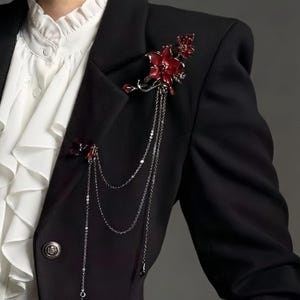 May include: A black blazer adorned with two red floral brooches. The brooches feature intricate details and dangle chains. The blazer is paired with a white ruffled shirt, creating a striking contrast.