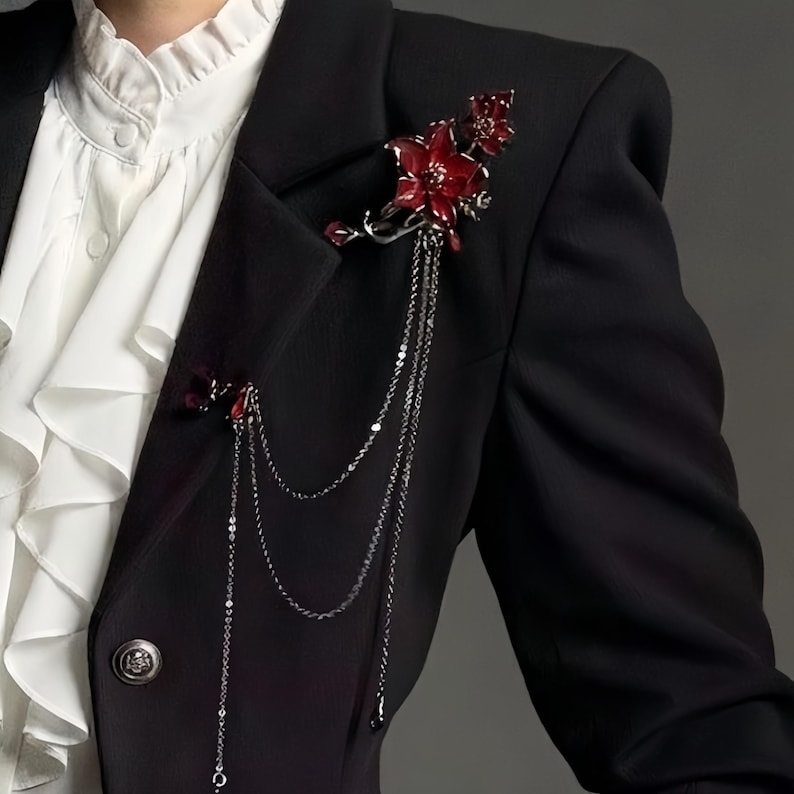 May include: A black blazer adorned with two red floral brooches. The brooches feature intricate details and dangle chains. The blazer is paired with a white ruffled shirt, creating a striking contrast.