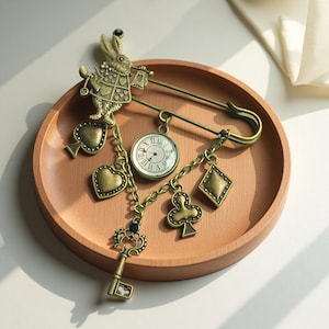 Op de afbeelding: Een vintage broche met een konijnfiguur, een zakhorloge en speelkaartsymbolen. De broche is gemaakt van antiek goudkleurig metaal en heeft een veiligheidsspeldsluiting. De bedels omvatten een hart, schoppen, klaveren en ruiten.