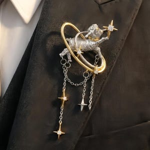 Puede incluir: Broche plateado y dorado con una figura de cabra dentro de un anillo dorado, adornado con cadenas colgantes y detalles en forma de estrella. El broche está sujeto a una blazer negra, un accesorio único de temática celestial.