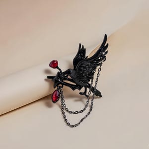May include: A black raven brooch with outstretched wings, perched on a branch. The raven holds a star with two red gemstone accents and dangling chains. The brooch is made of metal and has a gothic aesthetic.