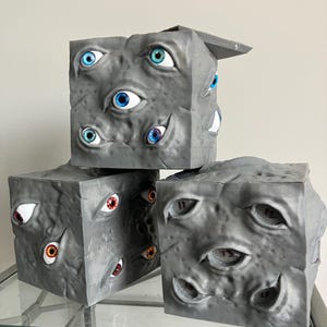 May include: Three gray, cube-shaped dice with multiple, varied-colored eyes embedded in their surfaces. The dice have a textured appearance and are arranged on a glass surface. One die has a lid open.