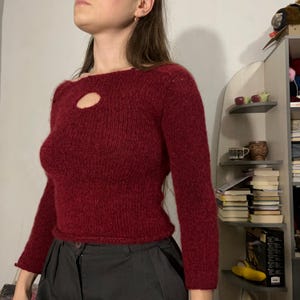 Könnte beinhalten: Ein weinroter Langarmpullover mit einem Schlüsselloch-Detail am Ausschnitt. Der Pullover hat einen gerippten Saum und Bündchen. Das Material scheint ein dicker Strick zu sein, der für kühleres Wetter geeignet ist. Der Pullover ist einfarbig, ohne sichtbare Muster.