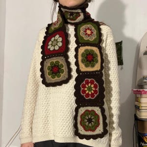 Może przedstawiać: Ręcznie robiony szalik z szydełka z powtarzającym się wzorem kwiatowym w kolorze czerwonym, zielonym, beżowym i brązowym. Szalik składa się z kwadratowych bloków z falistą krawędzią i jest noszony wokół szyi. Tło to kremowy sweter.