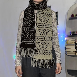 Puede incluir: Bufanda de punto con un diseño de mariposas. La bufanda es de dos tonos, con una sección marrón oscuro y una sección beige claro. El diseño de mariposa está en el color opuesto de cada sección. La bufanda tiene flecos en los extremos.