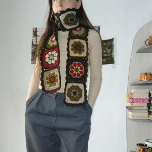 May include: A handmade crochet scarf and cowl set. The scarf features a series of square floral motifs in red, green, brown, and cream colors. The edges are scalloped. The cowl is attached to the scarf. The person is wearing a beige long-sleeve shirt and gray pants.