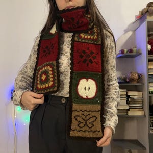May include: A patchwork scarf with a matching neck warmer. The scarf features squares of red, green, and brown with various designs, including an apple, a butterfly, and floral patterns. The scarf is made of a knitted material.