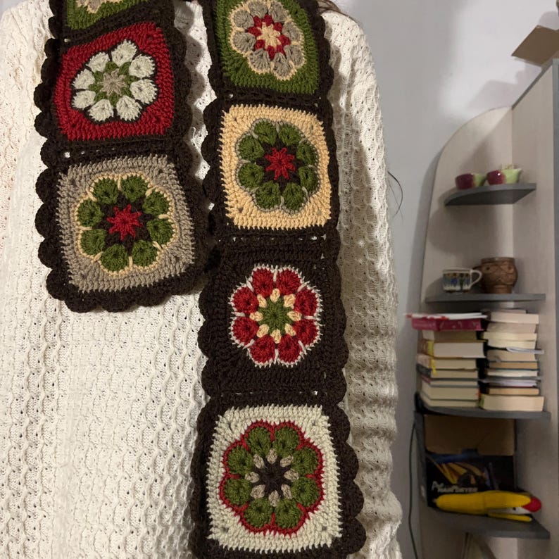 May include: A handmade crochet scarf featuring square floral motifs in red, green, beige, and brown. The scarf has a scalloped edge and is displayed against a white sweater. The scarf is approximately 60 inches long.