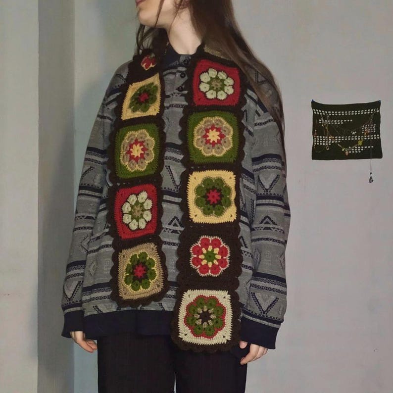 May include: A handmade crochet scarf featuring a repeating pattern of colorful floral squares. The scarf showcases a palette of brown, red, green, and yellow, creating a cozy and artistic accessory. The scarf is a unique, handcrafted item.