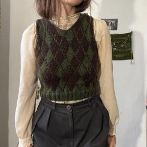 Peut inclure: Un gilet sans manches en tricot argyle vert et marron. Le pull sans manches a un col en V et un motif losange classique. Le gilet est porté sur un chemisier crème et associé à un pantalon gris foncé. L'ensemble est d'inspiration vintage.